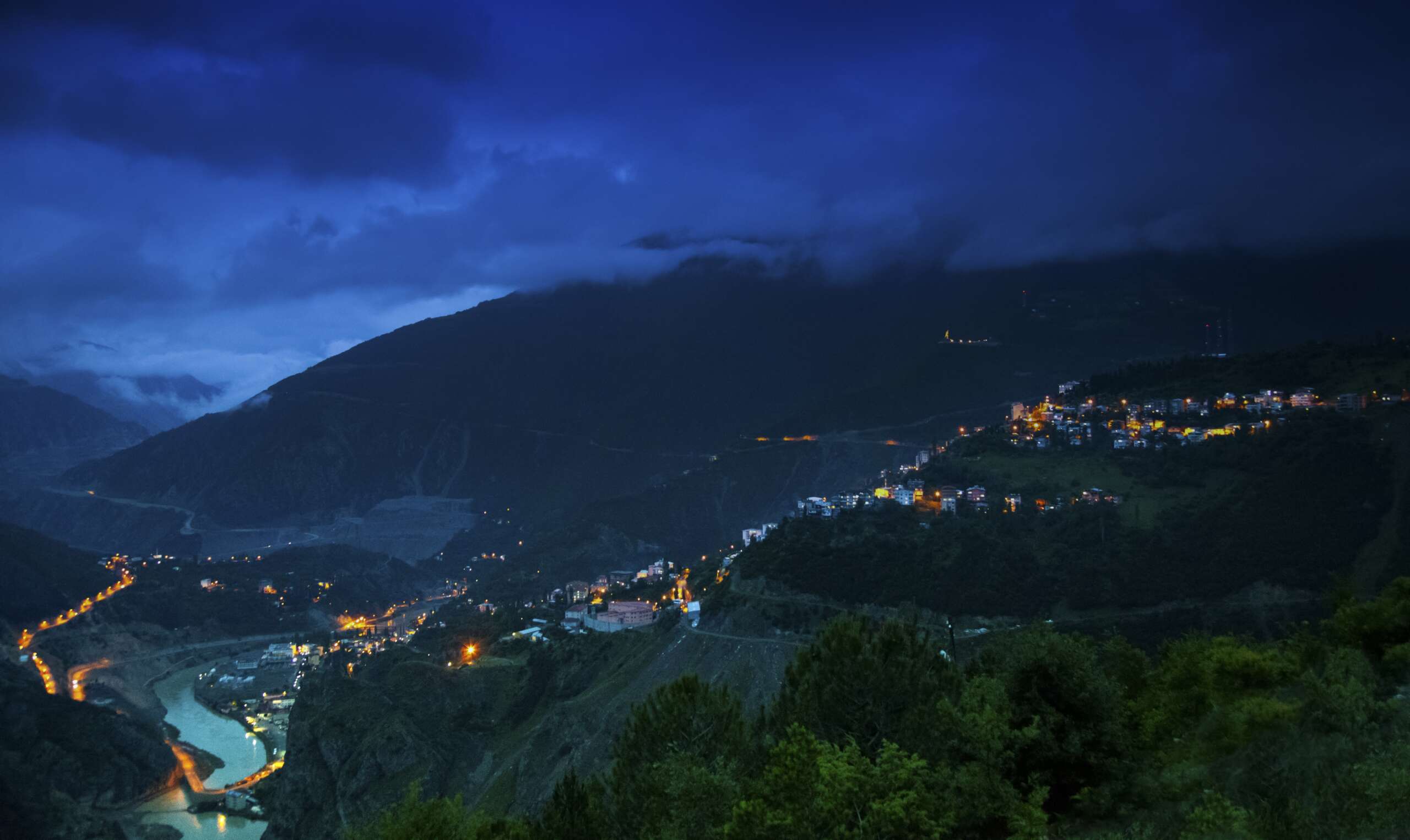 A landscape of hills covered in buildings and forests under a cloudy sky during the night
